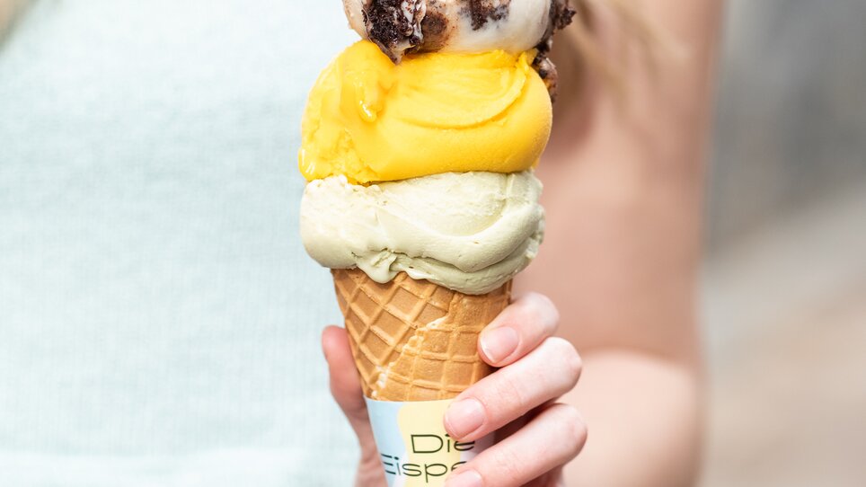 A young person holds a cone with three scoops of ice cream. | © Eisperle - Stiefkind Fotografie