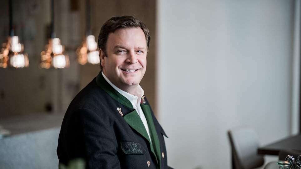 Ein Mann in traditioneller Kleidung lächelt am Tisch. Softe Beleuchtung im Hintergrund. | © Tina Szabo