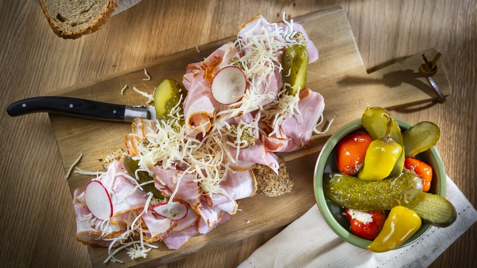 A wooden board topped with cold cuts, cheese, pickles, and a colorful antipasti side dish. | © Kaiser Greissler - Werner Krug