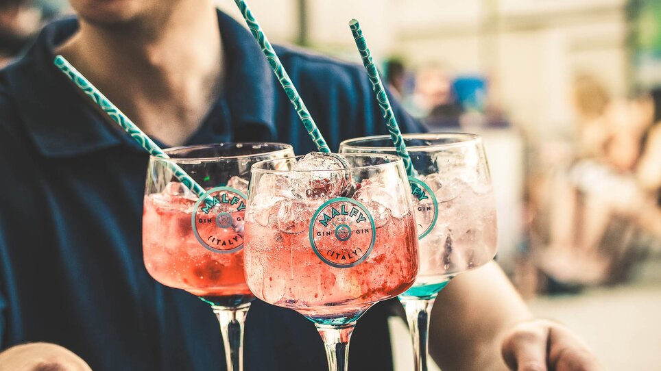 Ein fröhlicher Barkeeper serviert bunte Cocktails auf einem Tablett. | © Viertel 4