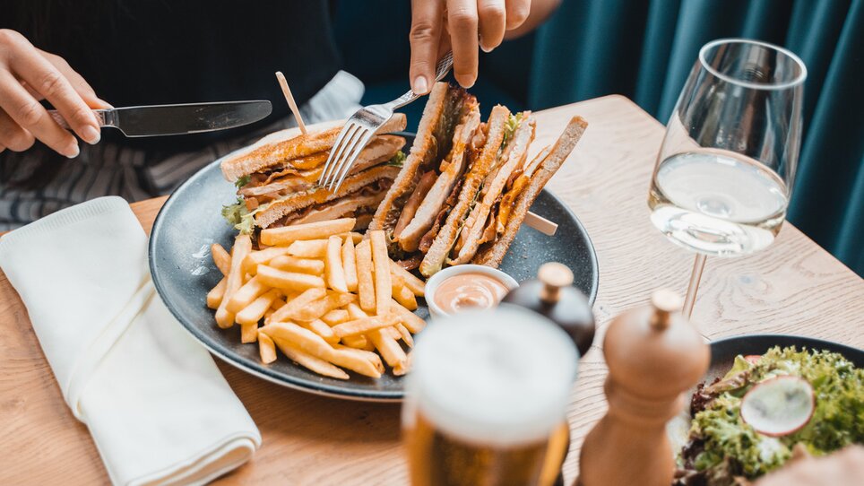A person in the restaurant is cutting a sandwich with chips on a plate, next to it is a glass of wine. | © Das Franz