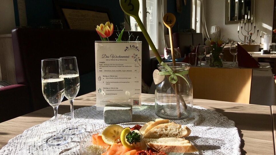 Ein Tisch im Restaurant mit Menükarte, zwei Gläsern Sekt und einer Platte mit Speisen. | © Das Lorenz