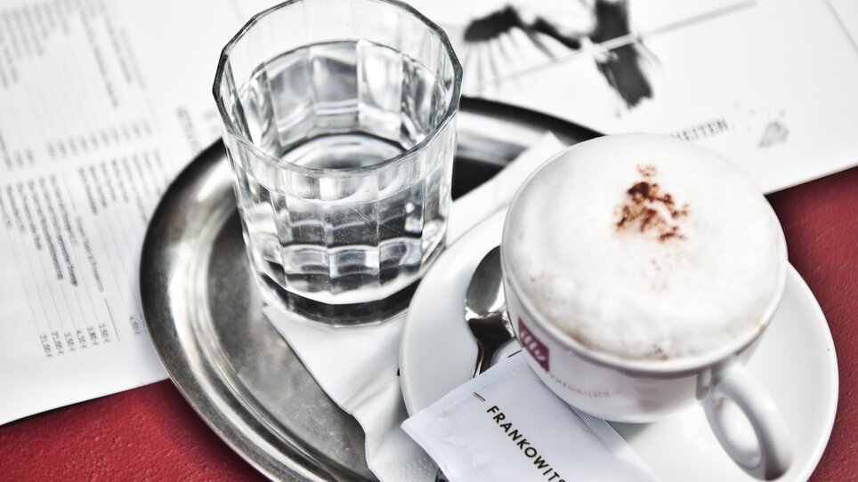 A cup of cappuccino and a glass of water with a sugar packet on a tray. | © Frankowitsch