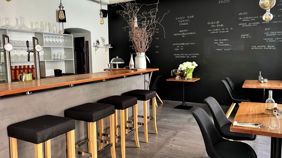 Un ristorante moderno con un bancone in legno, seggioline e una parete nera con le offerte di cibo e bevande. | © Gerüchteküche