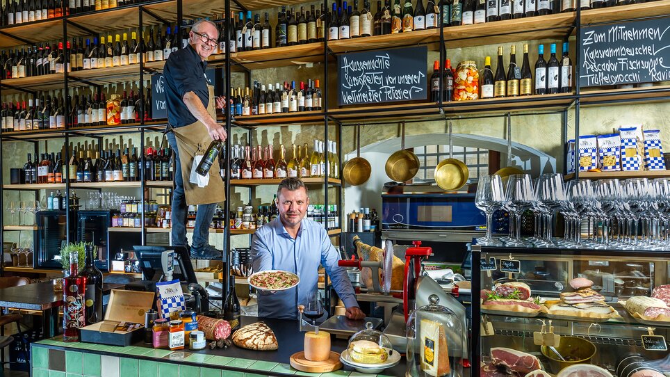 Zwei Männer in einem Restaurant mit Speisen und Getränken, einer serviert und der andere bereitet eine Platte vor. | © Paul Stajan