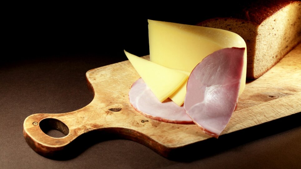 Slices of cheese and ham with bread on a wooden board. | © Sattler