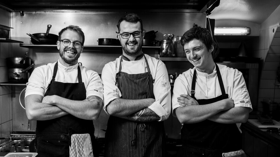 Three chefs smile in a kitchen with fresh fish.