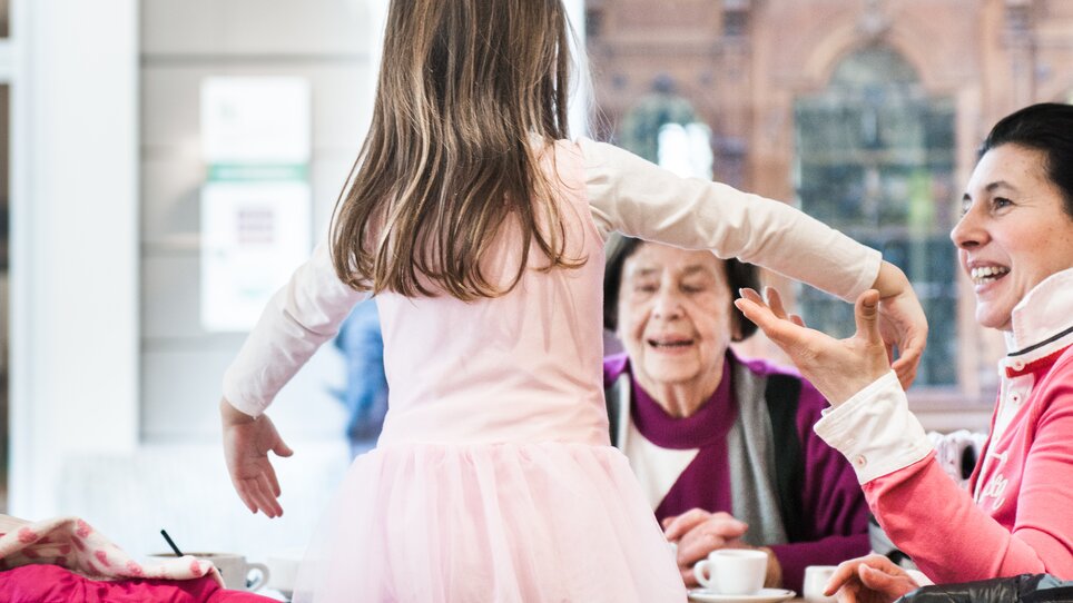 Ein Mädchen in einem rosa Kleid tanzt an einem Tisch mit fröhlichen Erwachsenen in einem Familien-Café in Graz. | © Martin Auer 