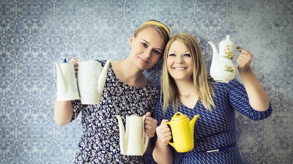 Zwei Frauen posieren mit verschiedenen Teekannen vor einer Tapete. | © Evgenia Rieger Photography 