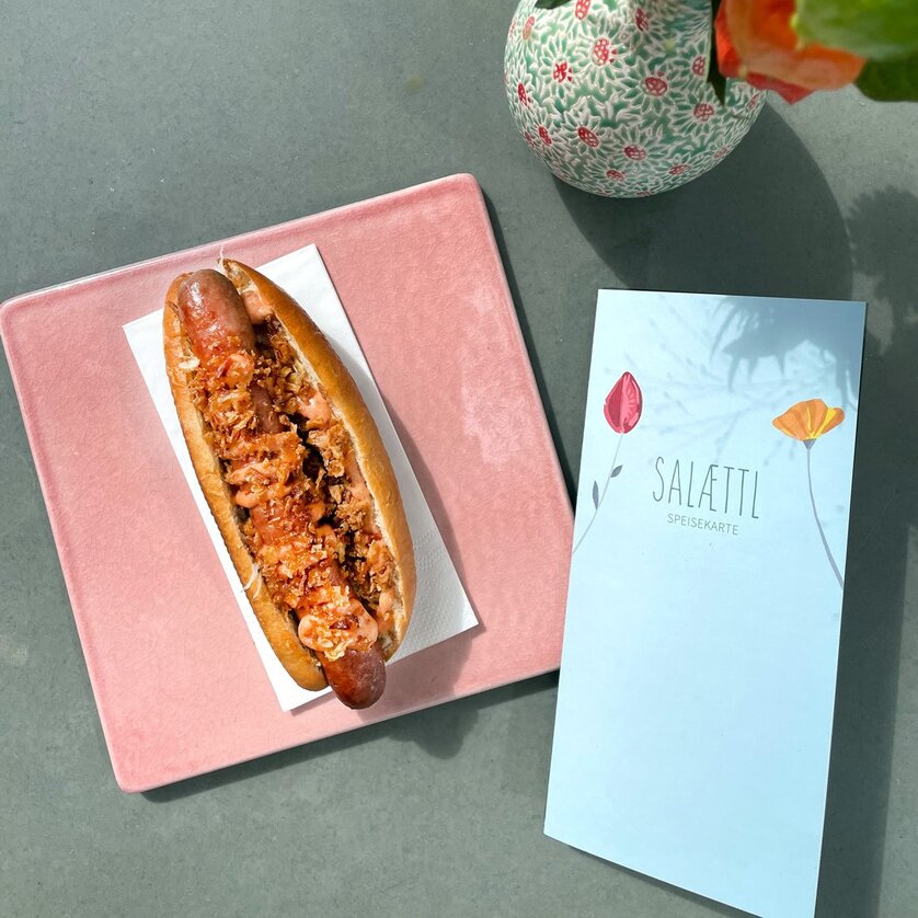 Ein Hotdog auf rosa Platte mit Speisekarte in einem modernen Café. | © Salaettl