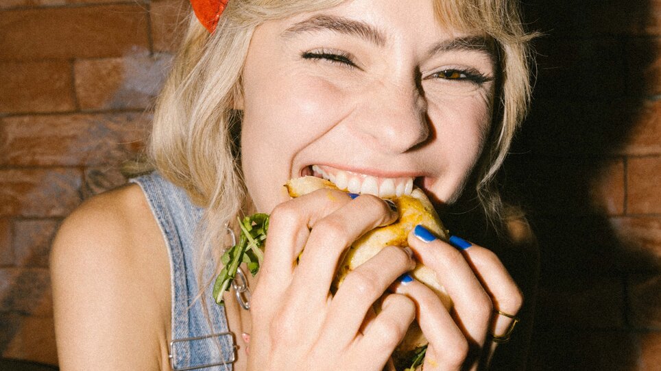 Una donna sorride e gusta un hamburger mentre sorride ampiamente. | © Bobby's Agency - Simon Bauer