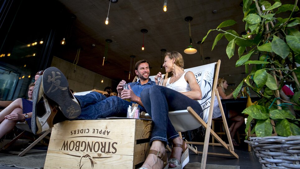 Two people are laughing and chatting in a cozy setting with plants and lighting. | © Graz Tourismus - Tom Lamm