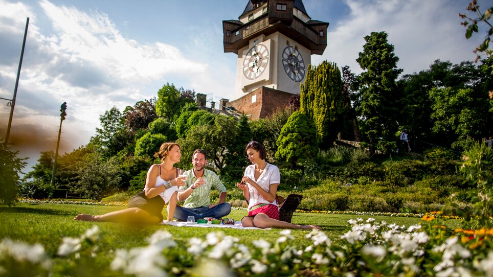Freunde genießen eine Auszeit am Schlossberg in Graz, mit Grazer Uhrturm im Hintergrund und picknicken gemeinsam. | © Graz Tourismus - Tom Lamm