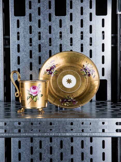 Ein goldener Becher und Untertasse mit Blumenmotiven auf einem Regal. | © Markus Mansi_MOMA