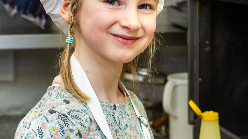 Ein Mädchen mit einer Kochmütze lächelt in einer Küche. | © Graz Tourismus - Paul Stajan
