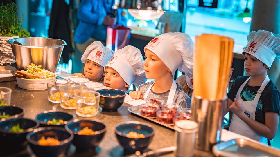 Kinder in Kochmützen bei einem Kochkurs in Graz, umgeben von Zutaten. | © KHC - Bernhard Schindler