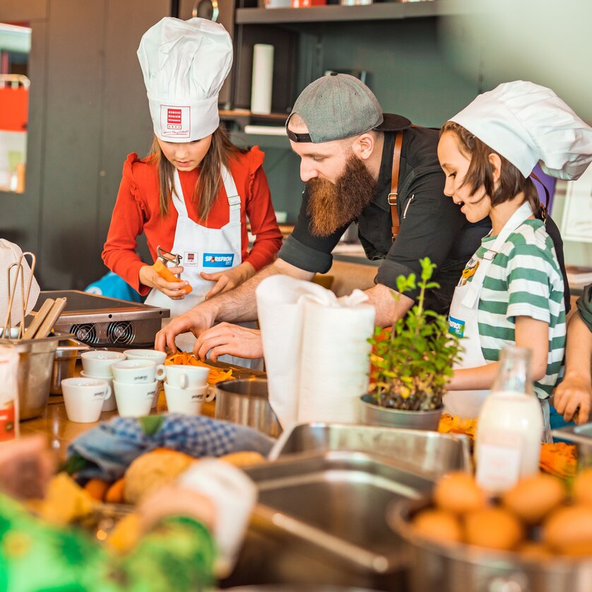 Kinder und ein Koch bereiten gemeinsam Gerichte in einer Kochschule vor. | © KHC - Bernhard Schindler