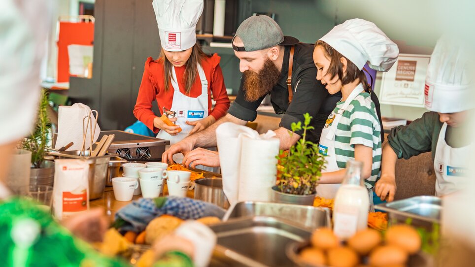 Kinder und ein Koch bereiten gemeinsam Gerichte in einer Kochschule vor. | © KHC - Bernhard Schindler