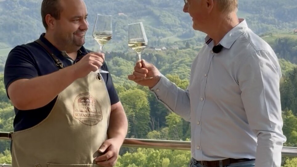 Two men clinking glasses, overlooking a green landscape. | © Schanzlwirt
