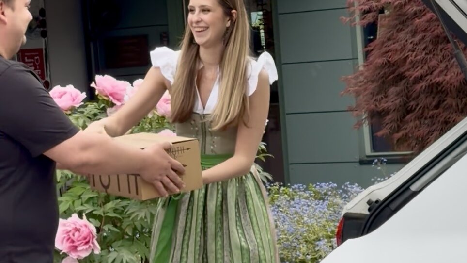 Woman in traditional dress hands a box to a man, surrounded by flowers. | © Schanzlwirt