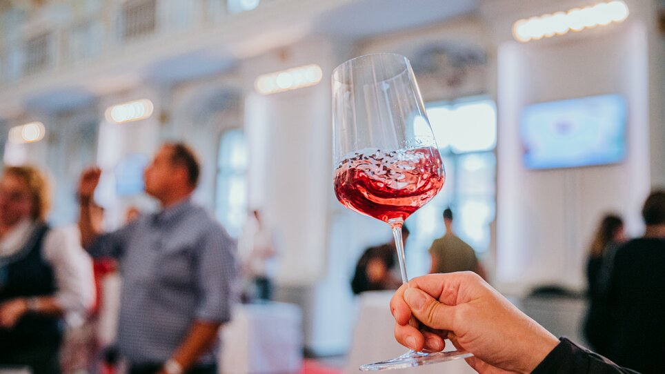 A wine glass with shimmering red wine is held up to the camera. The wine swirls in the glass, people can be recognised blurred in the background. The atmosphere is festive and stylish. | © Johanna Lamprecht