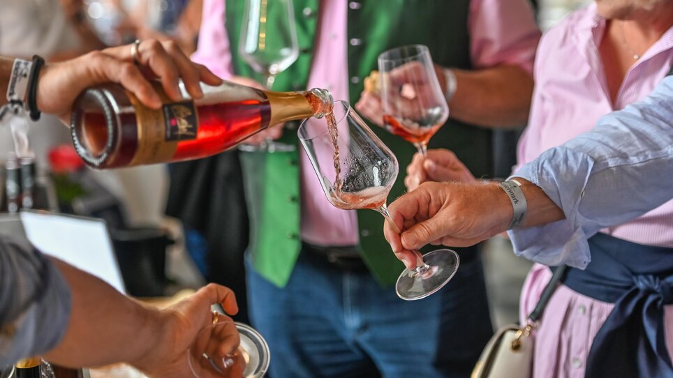 A rose-coloured sparkling wine is poured into a glass. The scene takes place in a convivial setting with festively dressed people - including a man in a green traditional waistcoat and a woman in a pink blouse. | © Wein Steiermark - Rosé Stainz