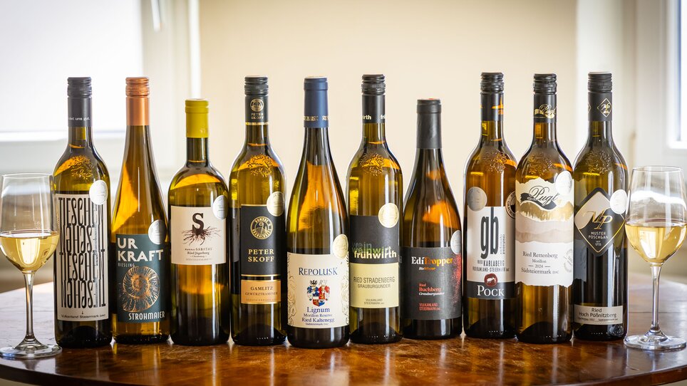 Eine Auswahl an Weißweinflaschen auf einem Holztisch, mit zwei Gläsern Wein. | © Graz Tourismus - Werner Krug