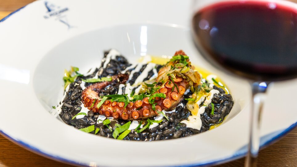 Un piatto di risotto nero con polpo e guarnizione, accompagnato da un bicchiere di vino rosso. | © Paul Stajan