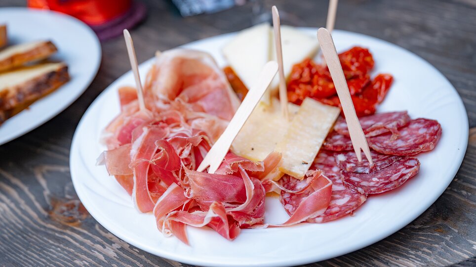 A plate with various types of meats, cheese, and sun-dried tomatoes, served with wooden sticks. | © 5komma5sinne - Rene Strasser