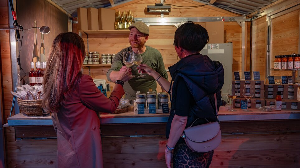 Two women clinking glasses, while a vendor showcases products in the background. | © 5komma5sinne - Rene Strasser