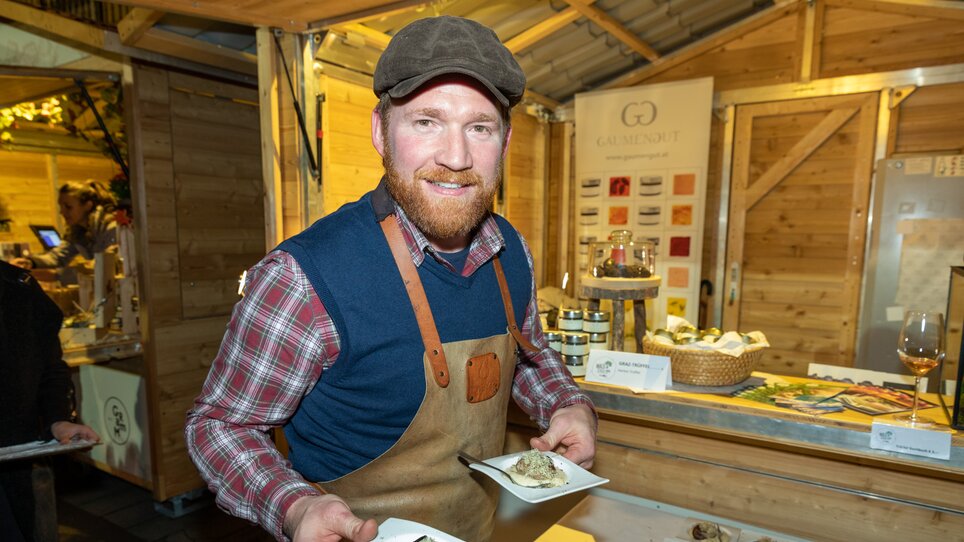 A smiling man in cooking attire holds two plates of truffle dishes at a festival. | © Graz Tourismus - Harry Schiffer