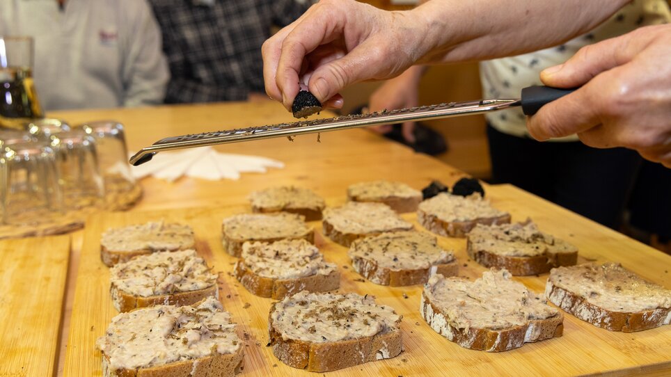 Eine Hand reibt frischen Trüffel über bestrichene Brote, im Hintergrund sind Personen zu sehen. | © Graz Tourismus - Harry Schiffer