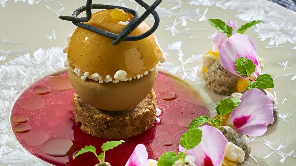 Un dessert artistico con una sfera di frutta su base di biscotto, circondato da salsa e decorazioni. | © Bastian Knapp