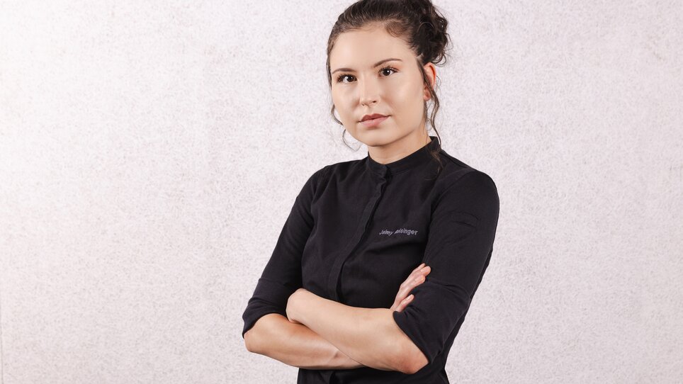 Selbstbewusst steht die junge Chef-Patissière Jaimy Reisinger mit verschränkten Armen in dunkler Kochkleidung. | © Pixelcoma
