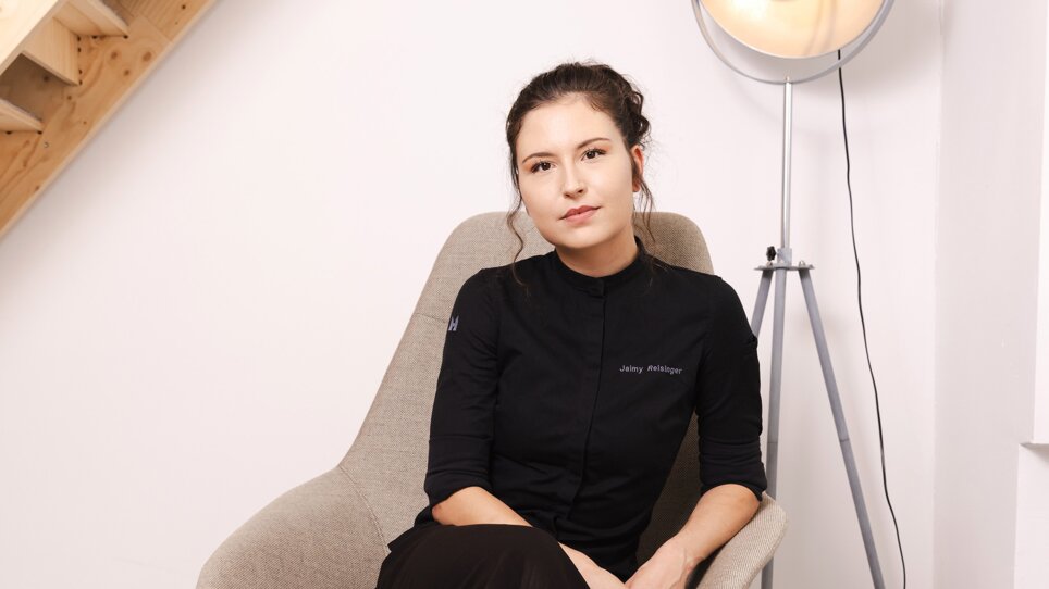 La chef pasticcera Jaimy Reisinger, vestita di nero, è seduta rilassata su una sedia grigia. | © Pixelcoma