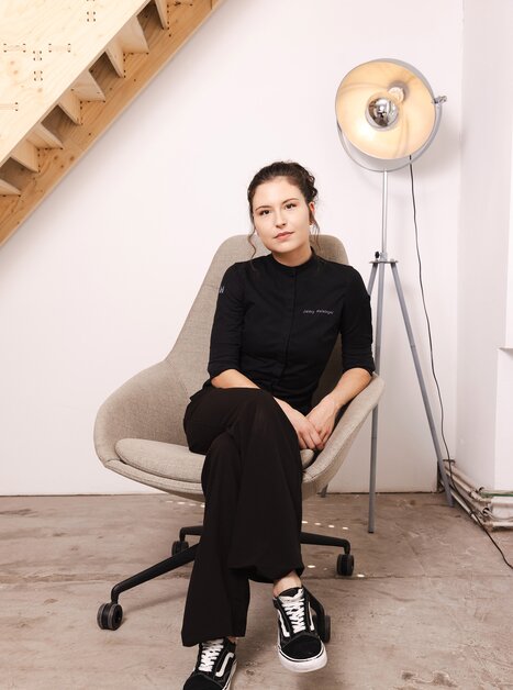 Chef-Patissière Jaimy Reisinger in schwarzem Outfit sitzt entspannt auf einem grauen Stuhl. | © Pixelcoma