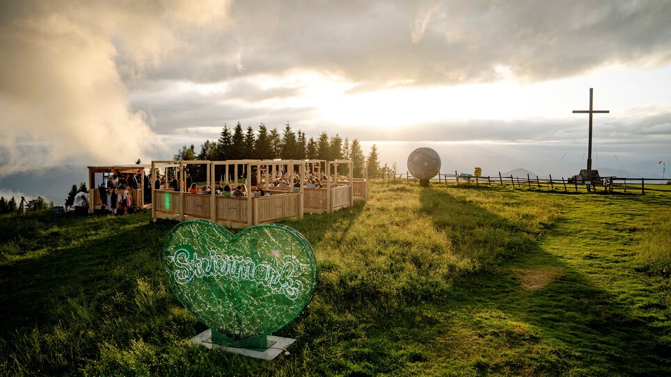 Eine Aussicht auf ein kulinarisches Event in den Bergen mit einem Herz-Symbol und einem Kreuz im Hintergrund. | © Region Graz - www
