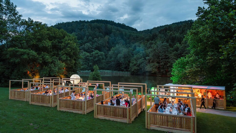 Eine wunderschöne Outdoor-Veranstaltung mit Holzbauten, beleuchtet von Lichtern, umgeben von Natur und einem Gewässer. | © Region Graz - www