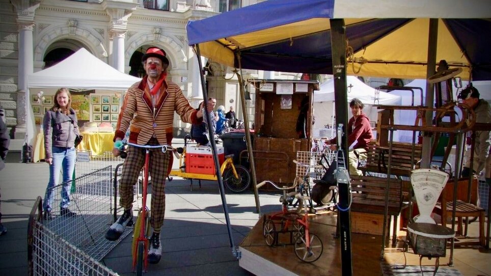 A clown on a bicycle at the Easter market in Graz, with a colorful tent and audience in the background. | © KIKH 1