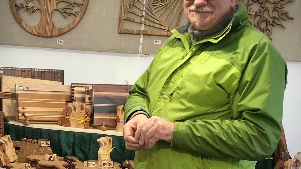 A man in a green jacket at a wooden market with wood crafts. | © KIKH 5