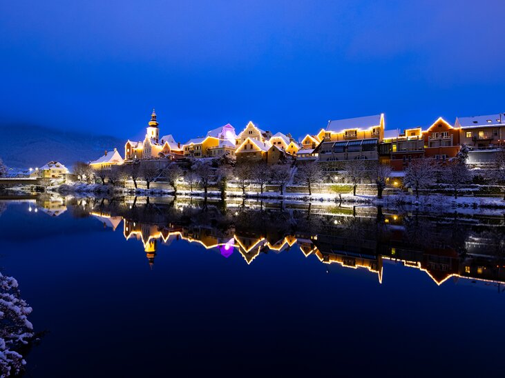 Beleuchtete Gebäude in Frohnleiten reflektieren im Wasser, umgeben von Schnee. | © Region Graz - Harry Schiffer