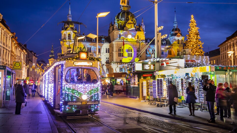 A festively lit Advent tram in Graz, surrounded by a winter scene and Christmas market. | © Graz Tourismus - Werner Krug