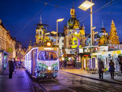 Die festlich beleuchtete Advent-Bim in Graz, umgeben von winterlicher Kulisse und Weihnachtsmarkt. | © Graz Tourismus - Werner Krug