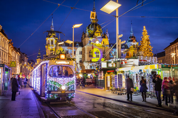 Die festlich beleuchtete Advent-Bim in Graz, umgeben von winterlicher Kulisse und Weihnachtsmarkt. | © Graz Tourismus - Werner Krug