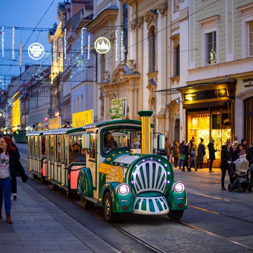A festive Advent train passes through the illuminated streets of Graz. People stroll along the shopping street. | © Graz Tourismus - Harry Schiffer