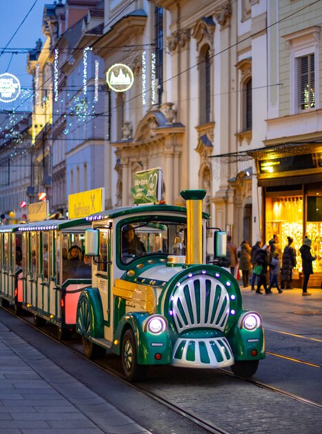 Ein festlicher Adventzug fährt durch die beleuchtete Grazer Herrengasse. Menschen schlendern entlang der Einkaufsstraße. | © Graz Tourismus - Harry Schiffer