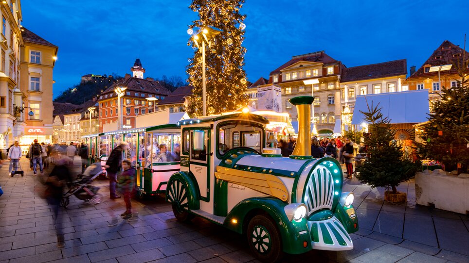 Eine festliche Stadtansicht von Graz zur Adventszeit mit einem beleuchteten Adventzug, der durch die Straßen fährt. | © Graz Tourismus-Harry Schiffer