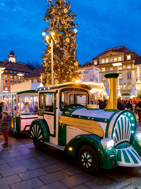 Eine festliche Stadtansicht von Graz zur Adventszeit mit einem beleuchteten Adventzug, der durch die Straßen fährt. | © Graz Tourismus-Harry Schiffer