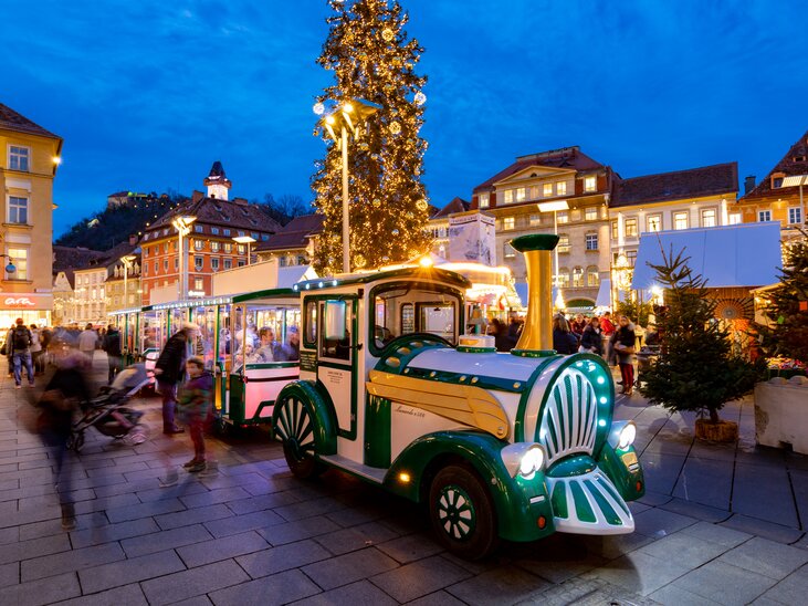 Eine festliche Stadtansicht von Graz zur Adventszeit mit einem beleuchteten Adventzug, der durch die Straßen fährt. | © Graz Tourismus-Harry Schiffer