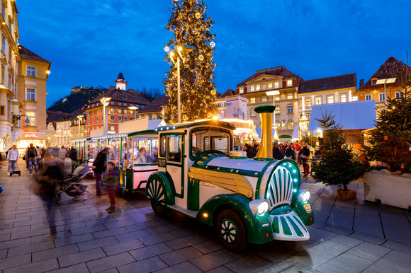 Eine festliche Stadtansicht von Graz zur Adventszeit mit einem beleuchteten Adventzug, der durch die Straßen fährt. | © Graz Tourismus-Harry Schiffer